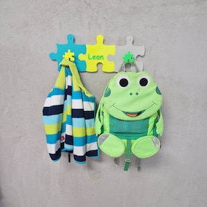 May include: A green frog-shaped backpack hangs on a colorful puzzle-shaped wall hook with the name "Leon" written on it. A blue and white striped jacket hangs on the hook next to the backpack.