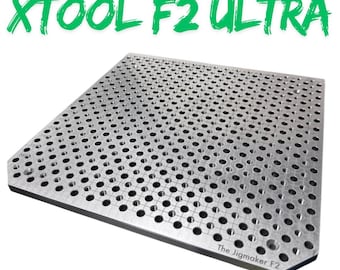 Puede incluir: Una placa de plantilla de metal plateado con numerosos agujeros espaciados uniformemente. El texto "THE JIGMAKER FOR XTOOL F2 ULTRA" está en verde y negro encima de la placa. El texto "The Jigmaker F2" está en la esquina inferior derecha.