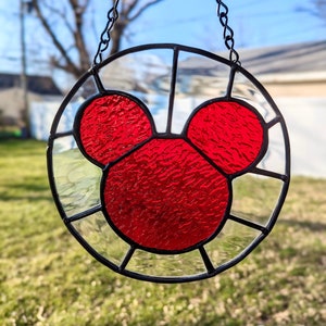 Może przedstawiać: Czerwona głowa Myszki Miki z witrażu, wisząca na czarnym łańcuchu. Witraż jest osadzony w okrągłej ramie z czarną obw&oacute;dką.