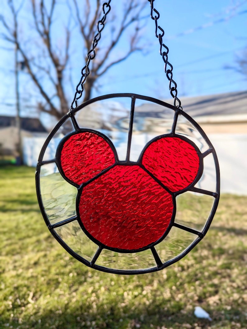 Może przedstawiać: Czerwona głowa Myszki Miki z witrażu, wisząca na czarnym łańcuchu. Witraż jest osadzony w okrągłej ramie z czarną obw&oacute;dką.