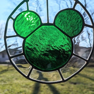 Może przedstawiać: Wisiorek z witrażu z zieloną sylwetką Myszki Miki. Szkło jest przezroczyste z czarną metalową ramą.