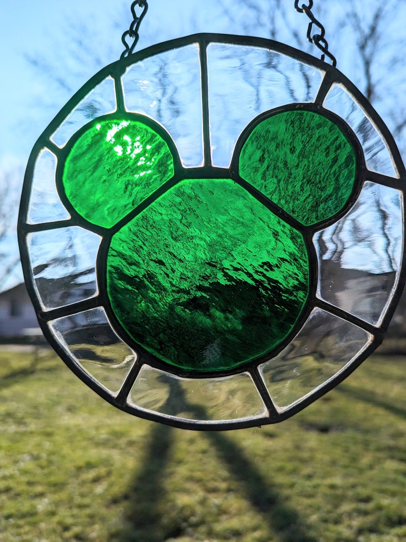 Może przedstawiać: Wisiorek z witrażu z zieloną sylwetką Myszki Miki. Szkło jest przezroczyste z czarną metalową ramą.