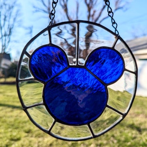 Może przedstawiać: Wiszące okno z witrażu z niebieską sylwetką Myszki Miki w środku. Okno jest otoczone przez przezroczyste panele szklane.