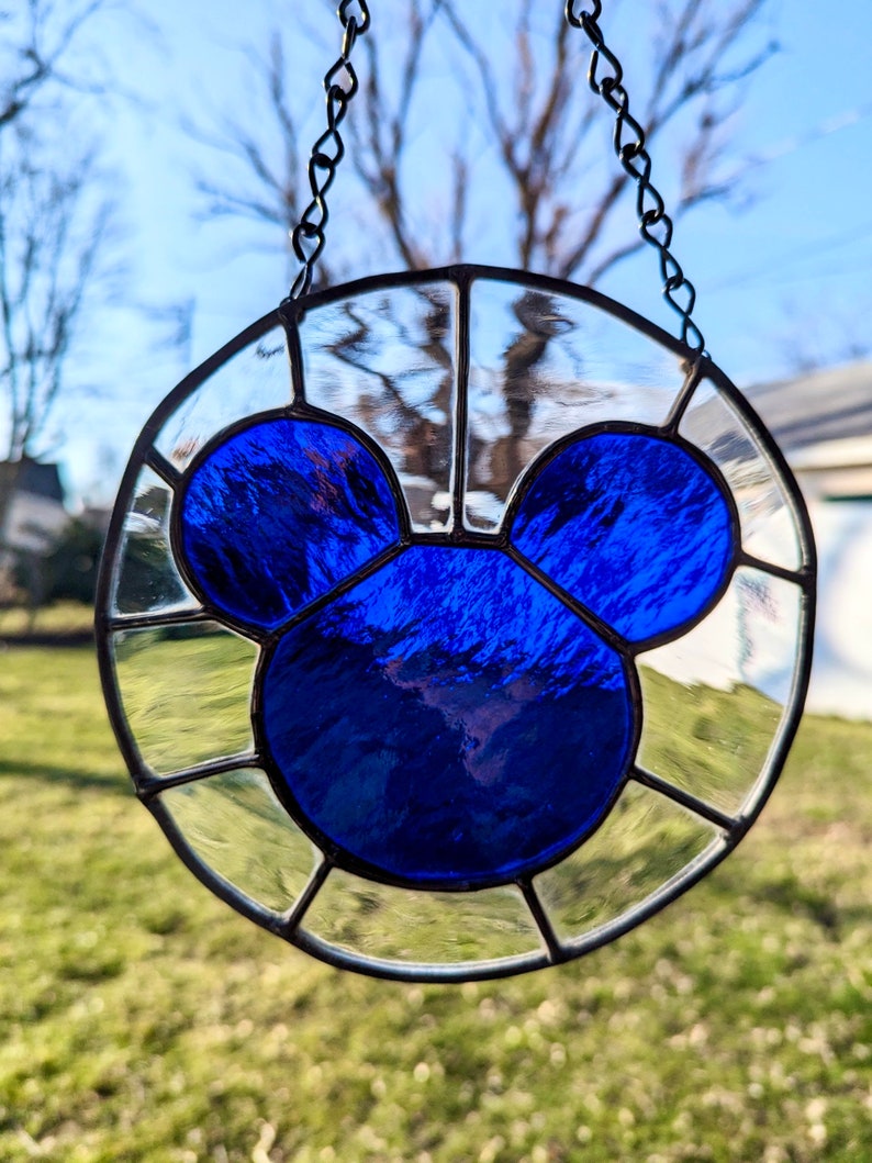 Może przedstawiać: Wiszące okno z witrażu z niebieską sylwetką Myszki Miki w środku. Okno jest otoczone przez przezroczyste panele szklane.