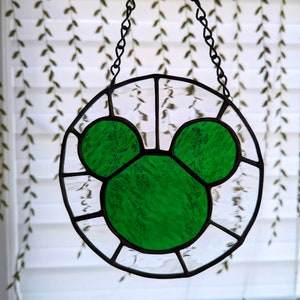 Może przedstawiać: Zielona głowa Myszki Miki z witrażu wisząca na łańcuchu. Szkło jest fakturowane i ma jasnozielony kolor.
