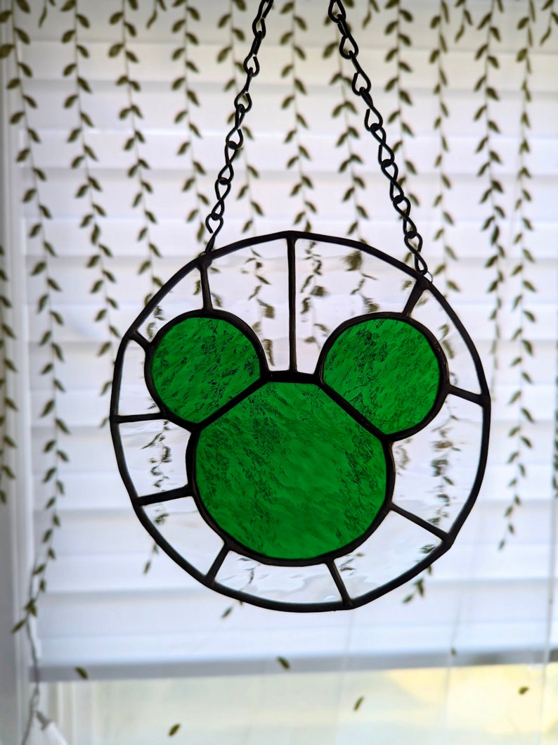 Może przedstawiać: Zielona głowa Myszki Miki z witrażu wisząca na łańcuchu. Szkło jest fakturowane i ma jasnozielony kolor.