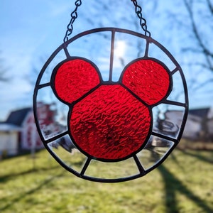 Może przedstawiać: Szklane okno w witrażu wiszące na czarnym łańcuchu. Okno jest okrągłe i przedstawia czerwony wz&oacute;r głowy Myszki Miki.