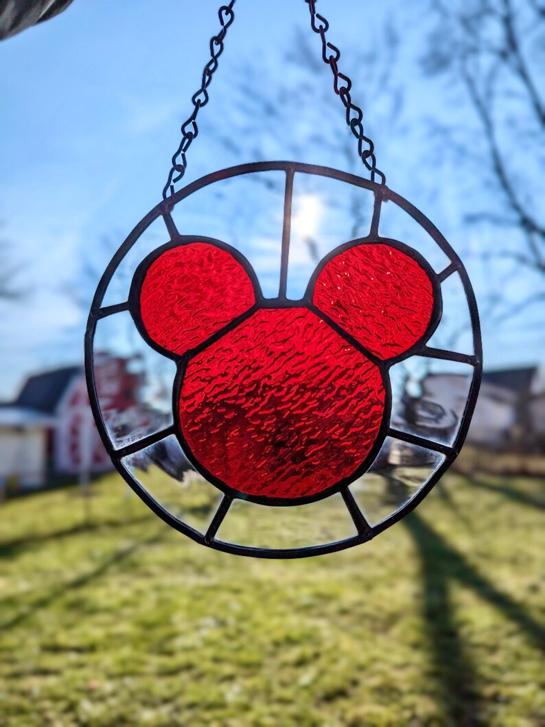 Może przedstawiać: Szklane okno w witrażu wiszące na czarnym łańcuchu. Okno jest okrągłe i przedstawia czerwony wz&oacute;r głowy Myszki Miki.