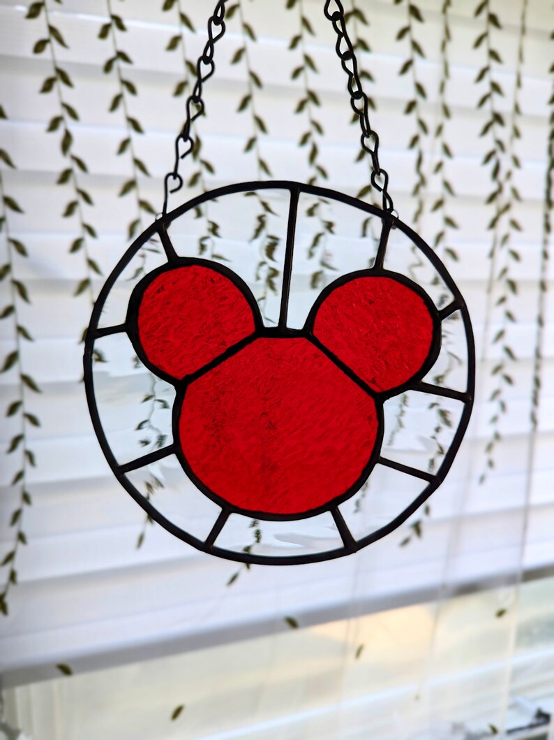 Może przedstawiać: Czerwona głowa Myszki Miki z witrażu wisząca na czarnym łańcuchu. Witraż jest osadzony w okrągłej ramie.