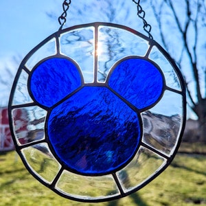 Może przedstawiać: Słoneczny łapacz wykonany ze szkła witrażowego z niebieską sylwetką Myszki Miki. Łapacz jest okrągły i ma łańcuszek do zawieszenia.