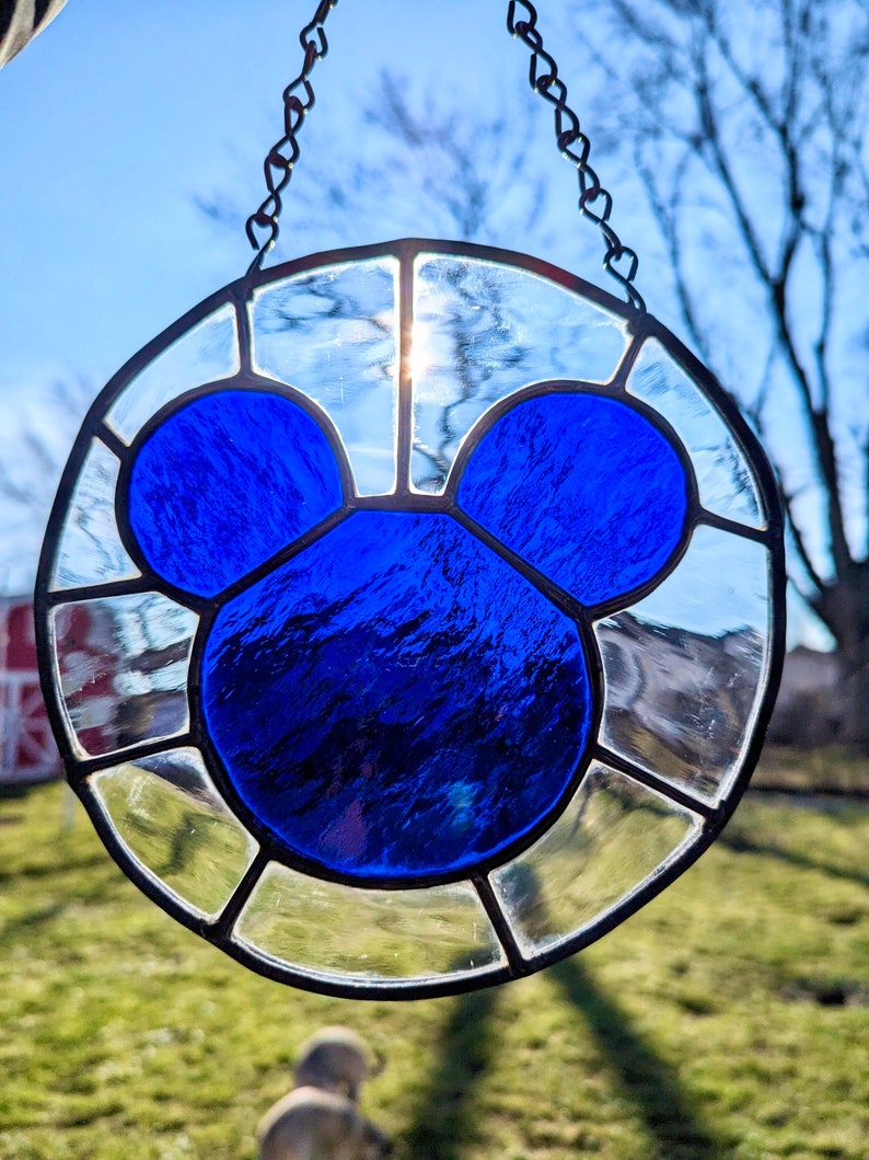 Może przedstawiać: Słoneczny łapacz wykonany ze szkła witrażowego z niebieską sylwetką Myszki Miki. Łapacz jest okrągły i ma łańcuszek do zawieszenia.