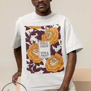 May include: White t-shirt featuring a floral design with large yellow roses and the text "TITLE FIGHT". The shirt is worn by a person wearing a white cap and tan shorts. The brand logo "Anarsha Apparel" is visible.
