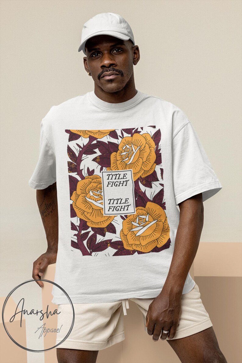 May include: White t-shirt featuring a floral design with large yellow roses and the text "TITLE FIGHT". The shirt is worn by a person wearing a white cap and tan shorts. The brand logo "Anarsha Apparel" is visible.