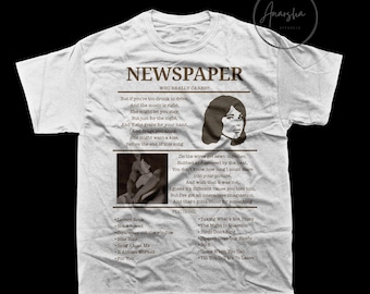 May include: White t-shirt with a newspaper-themed design. The shirt features a sepia-toned image of a woman and a couple, along with song titles and the word "NEWSPAPER". The shirt has a classic crew neck.