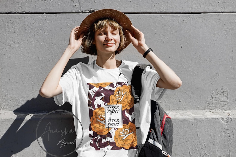 May include: White t-shirt with a floral design featuring orange roses and the text "TITLE FIGHT" in a rectangular box. The shirt is worn with a brown hat and a black backpack.