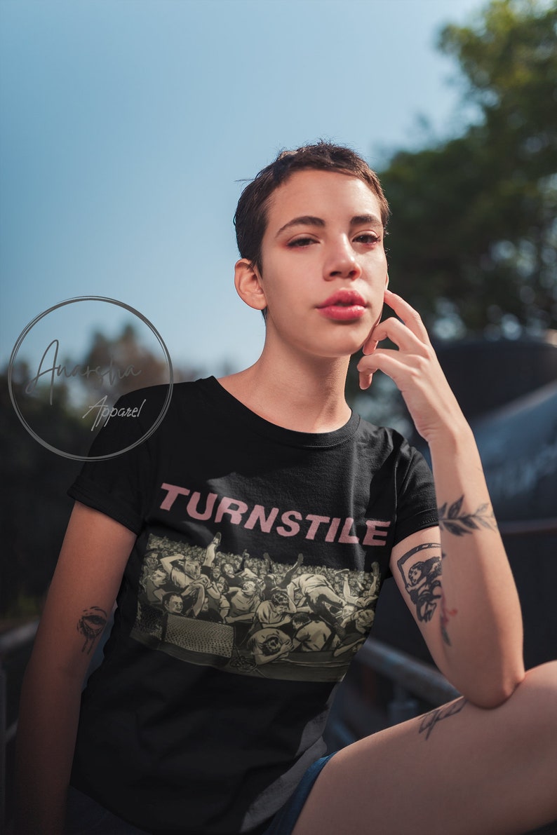Puede incluir: Una camiseta negra con un estampado gr&aacute;fico de una multitud de personas. El texto "TURNSTILE" est&aacute; impreso en rosa sobre el gr&aacute;fico.