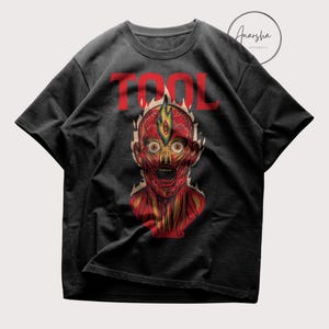 May include: Black t-shirt featuring the word "TOOL" in red, above a detailed illustration of a human head with exposed muscles and a fiery design. The graphic is in shades of red, yellow, and orange.