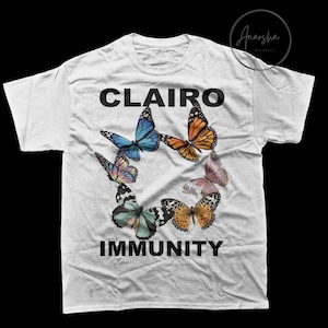 May include: White t-shirt featuring a colorful butterfly design arranged in a circle. The word "CLAIRO" is printed above the butterflies, and "IMMUNITY" is printed below. The butterflies are in shades of blue, orange, pink, and green.
