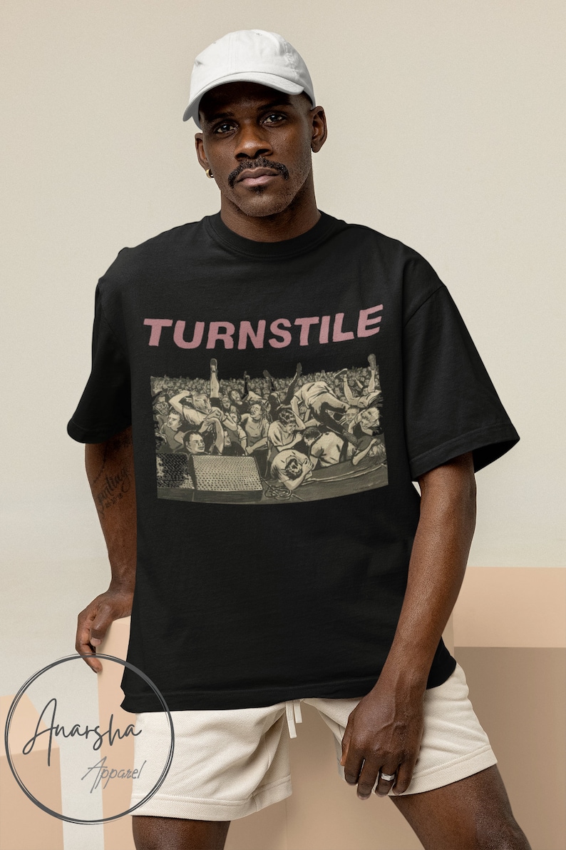 Puede incluir: Camiseta negra con un gr&aacute;fico de una multitud de personas en un concierto. El texto "TURNSTILE" est&aacute; impreso en rosa en la parte delantera de la camiseta.