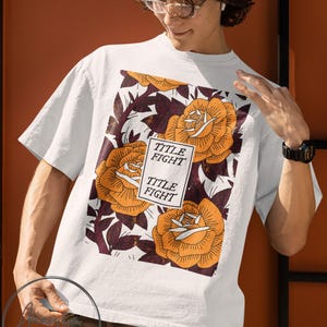 May include: White t-shirt featuring a floral design with orange roses and burgundy leaves. The shirt has the text "TITLE FIGHT" in a rectangular box. The shirt is a short-sleeved crew neck.