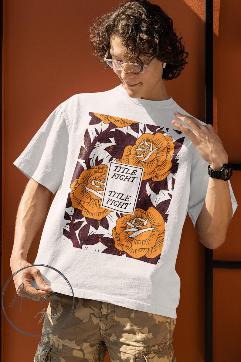 May include: White t-shirt featuring a floral design with orange roses and burgundy leaves. The shirt has the text "TITLE FIGHT" in a rectangular box. The shirt is a short-sleeved crew neck.