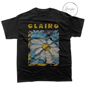 May include: Black t-shirt featuring the word "Clairo" in yellow block letters above a graphic of a daisy with a face. The daisy is white with a yellow center and is set against a blue background with yellow and black stripes.