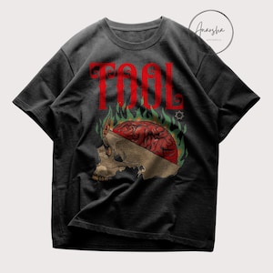 May include: Dark gray t-shirt featuring the word "TOOL" in red block letters above a skull graphic. The skull has a red brain and is surrounded by green and yellow flames. The shirt has short sleeves and a crew neck.