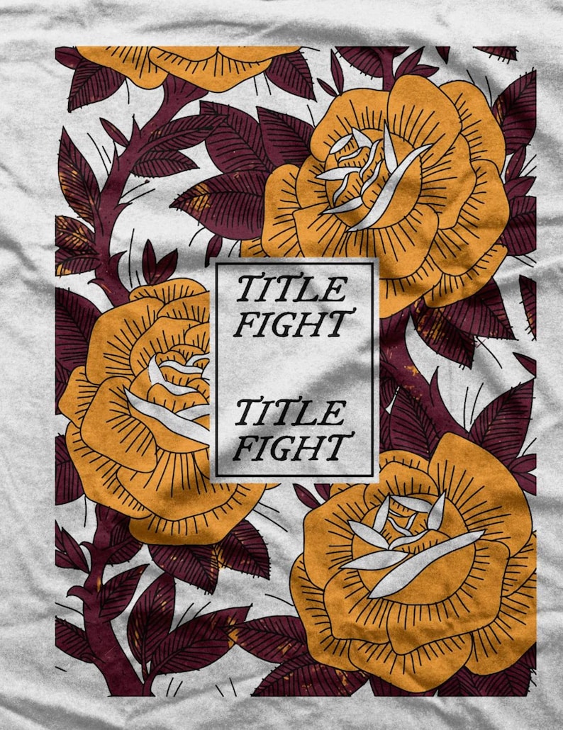 May include: A light gray t-shirt featuring a floral design with large yellow roses and burgundy leaves. The words "TITLE FIGHT" are printed in a white rectangle. The roses have detailed outlines.