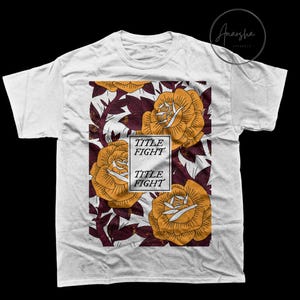 May include: White t-shirt featuring a floral design with large yellow roses and burgundy leaves. The design includes the text "TITLE FIGHT" in a white rectangular box. The shirt is a crew neck style.