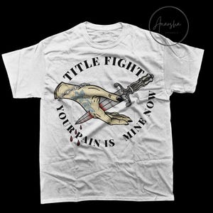 Peut inclure: T-shirt blanc avec un motif de style tatouage. Le design comprend une main tenant un poignard, avec le texte "TITLE FIGHT YOUR PAIN IS MINE NOW" disposé en cercle. Le graphisme est en beige, noir et rouge.