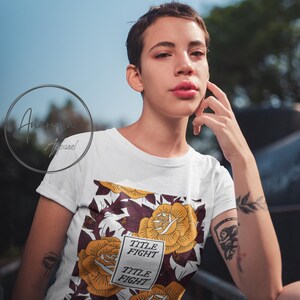 May include: White t-shirt featuring a floral design with yellow roses and burgundy leaves. The shirt includes the text "TITLE FIGHT" in a rectangular box. The design is set against a white background.