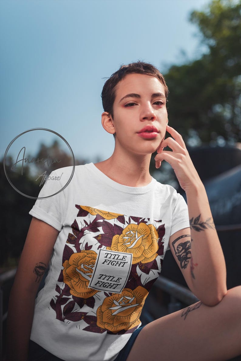 May include: White t-shirt featuring a floral design with yellow roses and burgundy leaves. The shirt includes the text "TITLE FIGHT" in a rectangular box. The design is set against a white background.