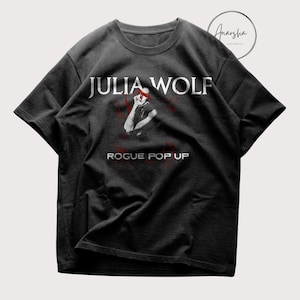 Puede incluir: Camiseta gris oscuro con el texto blanco "JULIA WOLF" y un gráfico de una persona. Debajo, el texto rojo dice "ROGUE POP UP". La camiseta tiene cuello redondo y mangas cortas.