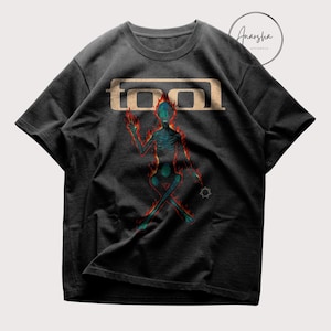 May include: Black t-shirt featuring a stylized figure with a fiery outline and a teal interior. The word "TOOL" is printed in a tan, blocky font above the figure. The shirt has short sleeves and a crew neck.