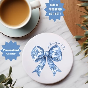 May include: A white ceramic coaster with a blue floral bow design and the name "Abigail" in cursive. The coaster is labeled "Personalized Coaster" and "Can be purchased as a set!" The text "Each coaster is sold separately" is also visible.