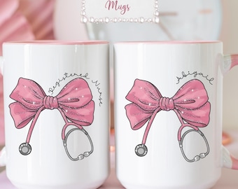 May include: Two white 15 oz ceramic mugs with pink handles and interiors. Each mug features a pink glittery bow graphic integrated with a stethoscope. One mug displays the text Registered Nurse, and the other displays the name Abigail in script.