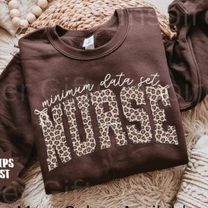 May include: A dark chocolate-colored sweatshirt with the words "minimum data set NURSE" printed on the front. The word "NURSE" is in a leopard print pattern. The text "More Colors Available" is at the bottom.