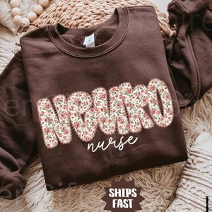 May include: A dark chocolate-colored sweatshirt with the word "NEURO" in large, floral-patterned letters and the word "nurse" in a cursive font. The sweatshirt has long sleeves and a crew neck. The text "SHIPS FAST" is also visible.