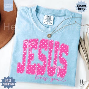 May include: A light blue short-sleeve t-shirt with a crew neckline. The shirt features the word JESUS in large, pink, white-polka-dotted letters with pink bows on the first and last letters, and the text loves you in pink cursive script below.