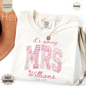 以下が含まれることがあります： アイボリー色のTシャツ。ピンク色の文字で「it's giving MRS Williams Est. 2026」と書かれています。「MRS」の文字は花柄とギンガムチェック柄でアップリケされています。金のネックレスとペンダントがシャツの上にあります。