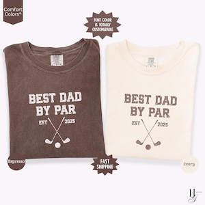 May include: Two t-shirts with a golf-themed design. The brown t-shirt says "Best Dad By Par" with a golf club and ball graphic. The ivory t-shirt says the same thing. Both shirts have "EST 2025" printed on them.