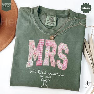 May include: A moss green t-shirt with the word "MRS" appliqued in pink floral and gingham fabric. The name "Williams" and the date "Est. 2026" are embroidered below. A gold necklace with a pendant rests on the shirt.