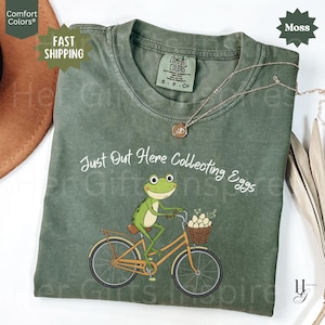 以下が含まれることがあります： カエルが自転車に乗り、カゴに卵を運んでいるモスグリーンのTシャツ。「Just Out Here Collecting Eggs」の文字がプリントされています。金のネックレスがシャツの上にかけられています。
