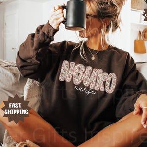 Puede incluir: Una sudadera de color chocolate oscuro con la palabra "NEURO" en letras grandes con estampado floral y la palabra "nurse" en una fuente cursiva m&aacute;s peque&ntilde;a. La sudadera es de manga larga y cuello redondo.