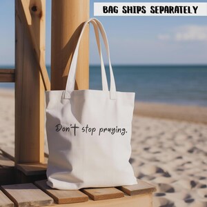 May include: Off-white canvas tote bag with long handles. The bag features the text "Don't stop praying" in a black script. The background includes a wooden structure, sand, and the ocean.