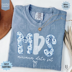 Può includere: Una maglietta blu jeans Comfort Colors con le lettere "MDS" in un motivo floreale e design a quadretti. Le parole "minimum data set" sono stampate sotto. Una collana d'oro con un ciondolo è drappeggiata sulla maglietta.