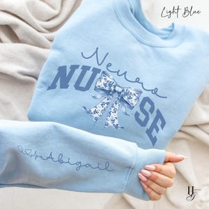 Könnte beinhalten: Hellblauer Pullover mit dem Text "Never Nurse" in blauer Schrift mit einer blauen und weißen Blumen Schleife. Der Pullover hat ein Herz mit einer Herzschlaglinie und den Namen "Abigail" auf dem Ärmel.