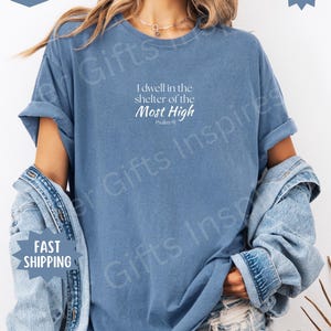 May include: A blue jean colored short-sleeve t-shirt featuring white text that reads I dwell in the shelter of the Most High Psalm 91. The shirt has a crew neckline and a relaxed fit, layered with a denim jacket over the shoulders.