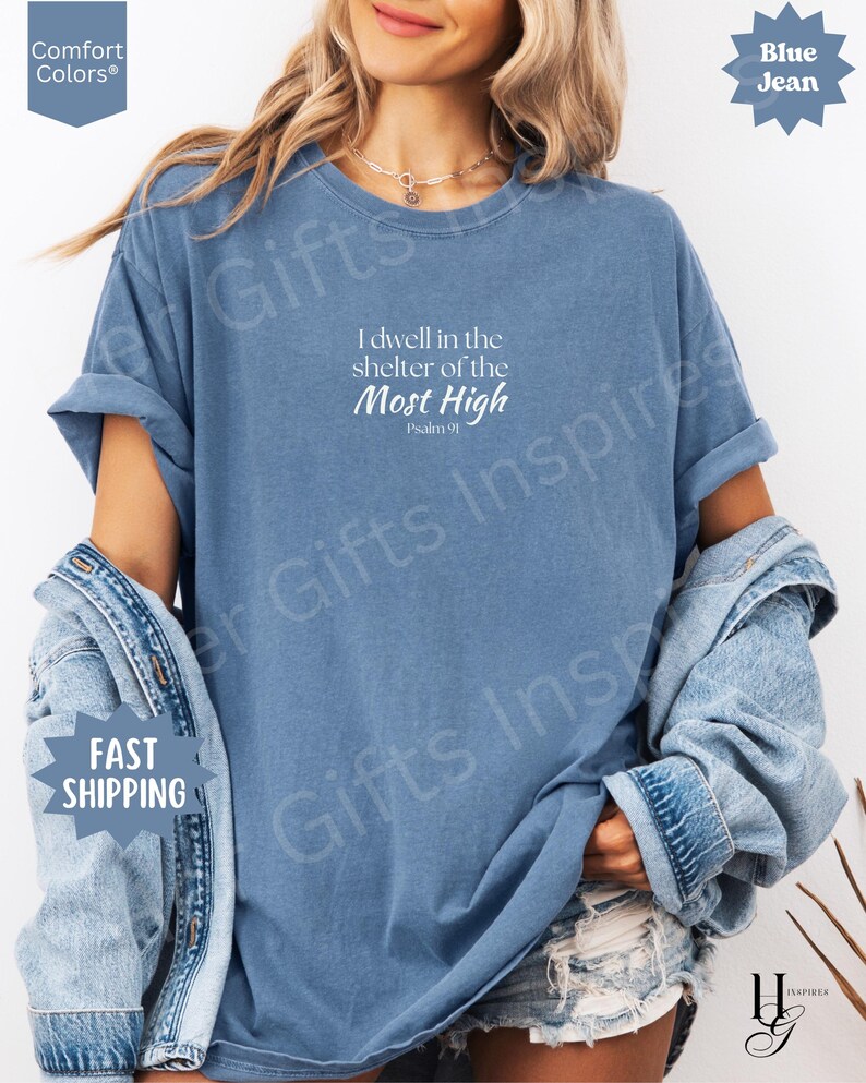 May include: A blue jean colored short-sleeve t-shirt featuring white text that reads I dwell in the shelter of the Most High Psalm 91. The shirt has a crew neckline and a relaxed fit, layered with a denim jacket over the shoulders.
