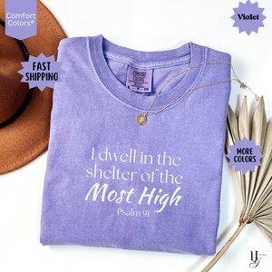May include: A violet short-sleeved t-shirt with a crew neckline and a Comfort Colors brand label. The front features white text that reads, I dwell in the shelter of the Most High, Psalm 91. A thin gold chain necklace with a small circular pendant rests on top.